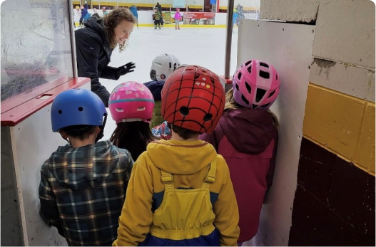 A group of child skaters in helmets listening to an instuctor and about to enter an ice rink